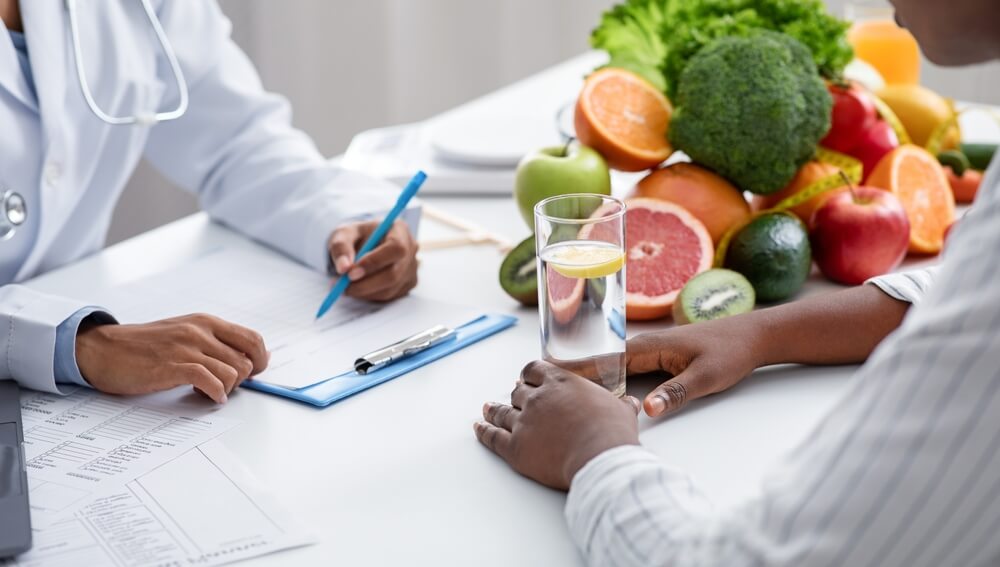 A doctor speaking with a patient, with a spread of vegetables on the desk. Learn more about detox from drugs as part of addiction treatment, here.